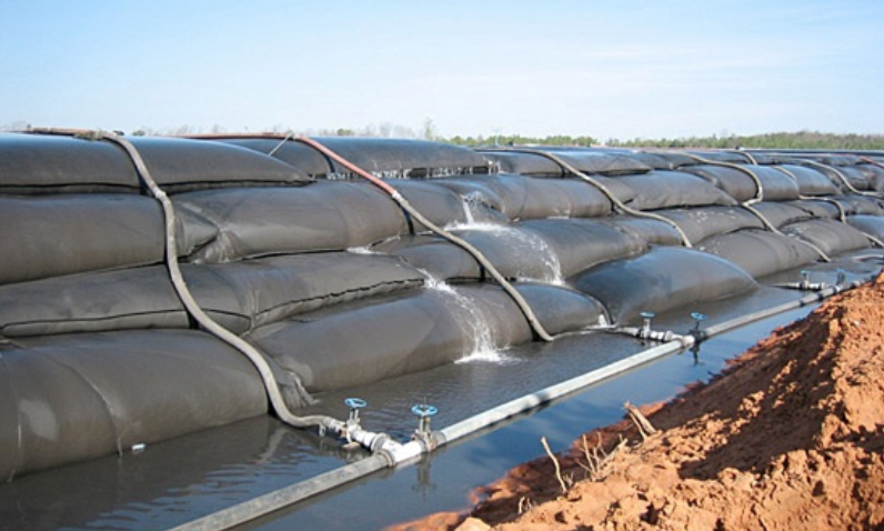 A row of black bags filled with water are sitting on top of a dirt field.