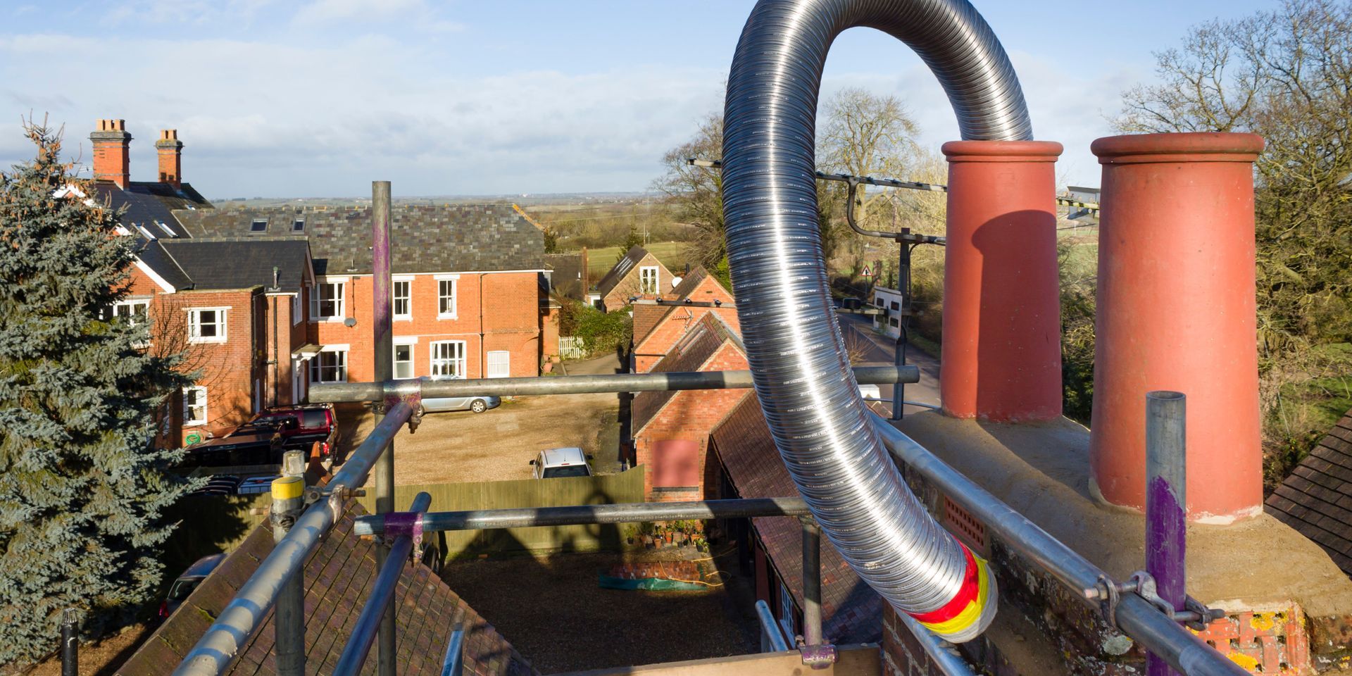 A curved metal flue pipe extends from the red chimney of a brick building, with scaffolding in the foreground.