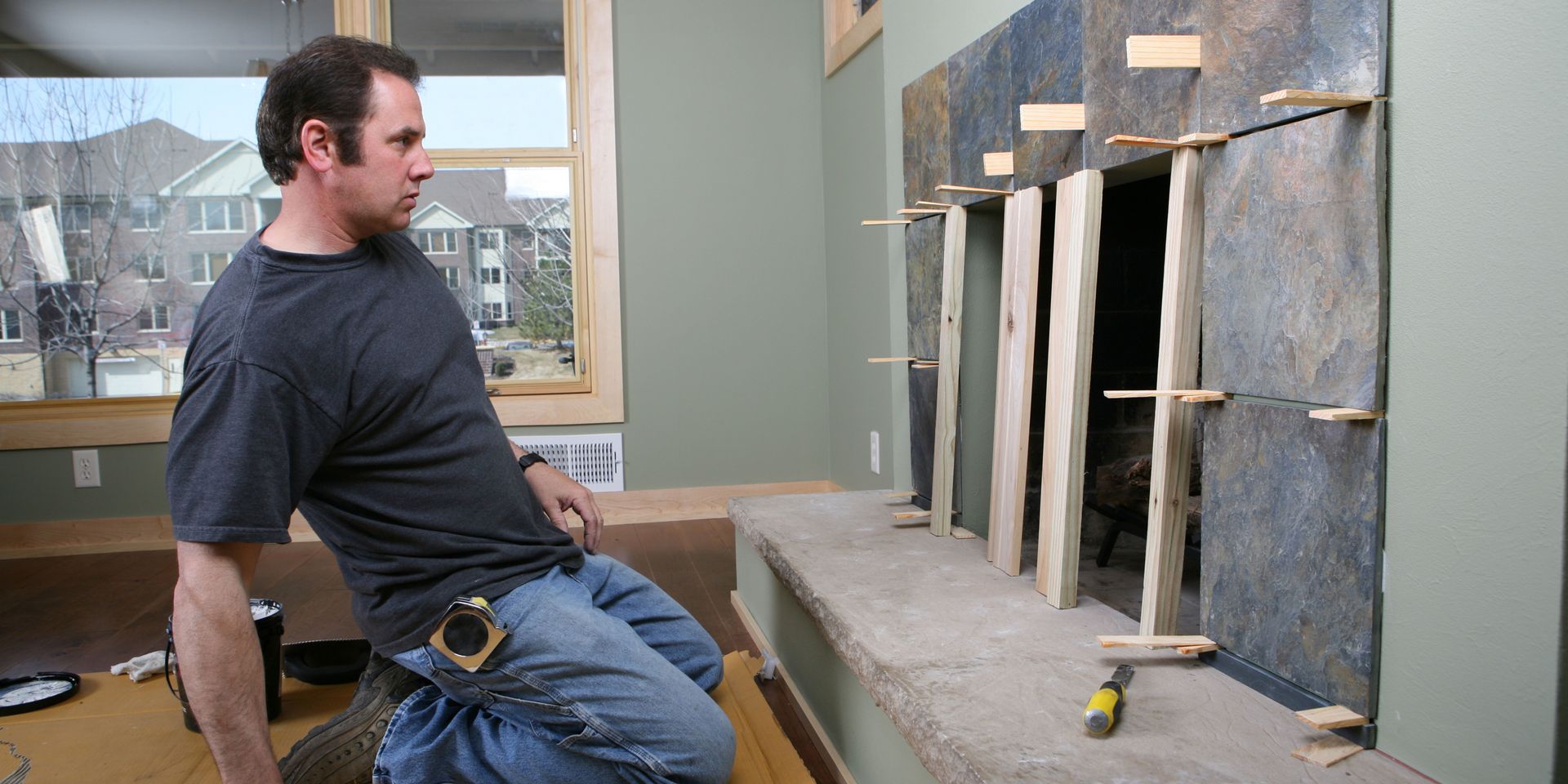 Man working on fireplace; setting tiles with wooden supports. Indoors, neutral wall color, and overcast day.