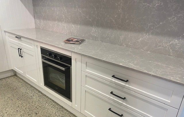 White kitchen cabinets with black handles, built-in oven, and light gray countertop and backsplash. — Kitchens Factory Direct in Port Macquarie, NSW