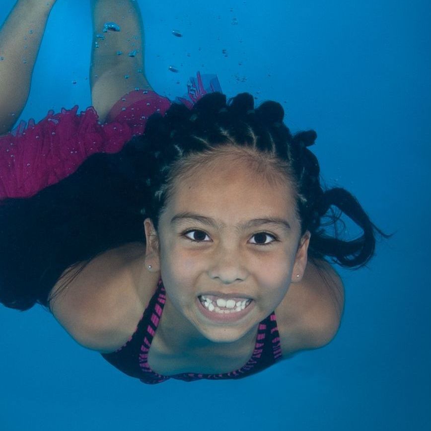 a little girl is swimming underwater in a pool and smiling during swim lessons.