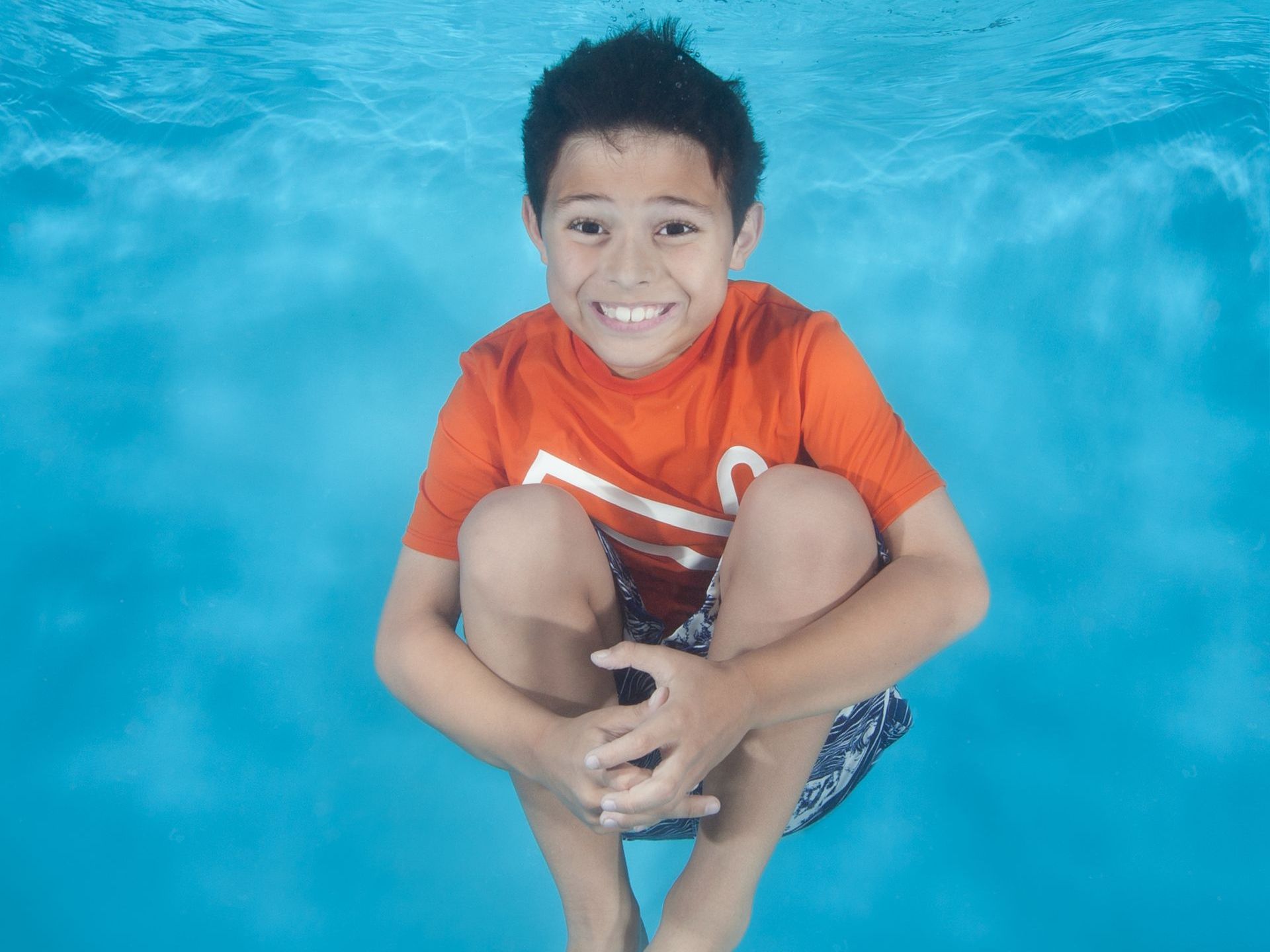 Un niño está sentado bajo el agua en una piscina y sonriendo, en clases de natación en Ocaquatics