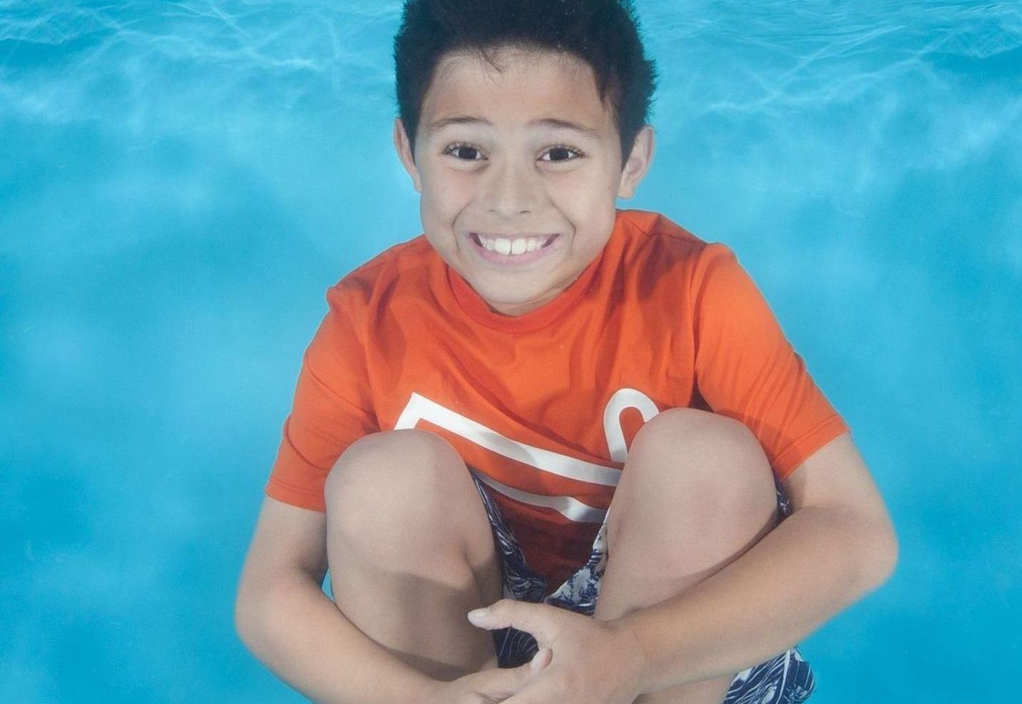 a young boy in an orange shirt is kneeling in a pool and smiling underwater in swim lessons .