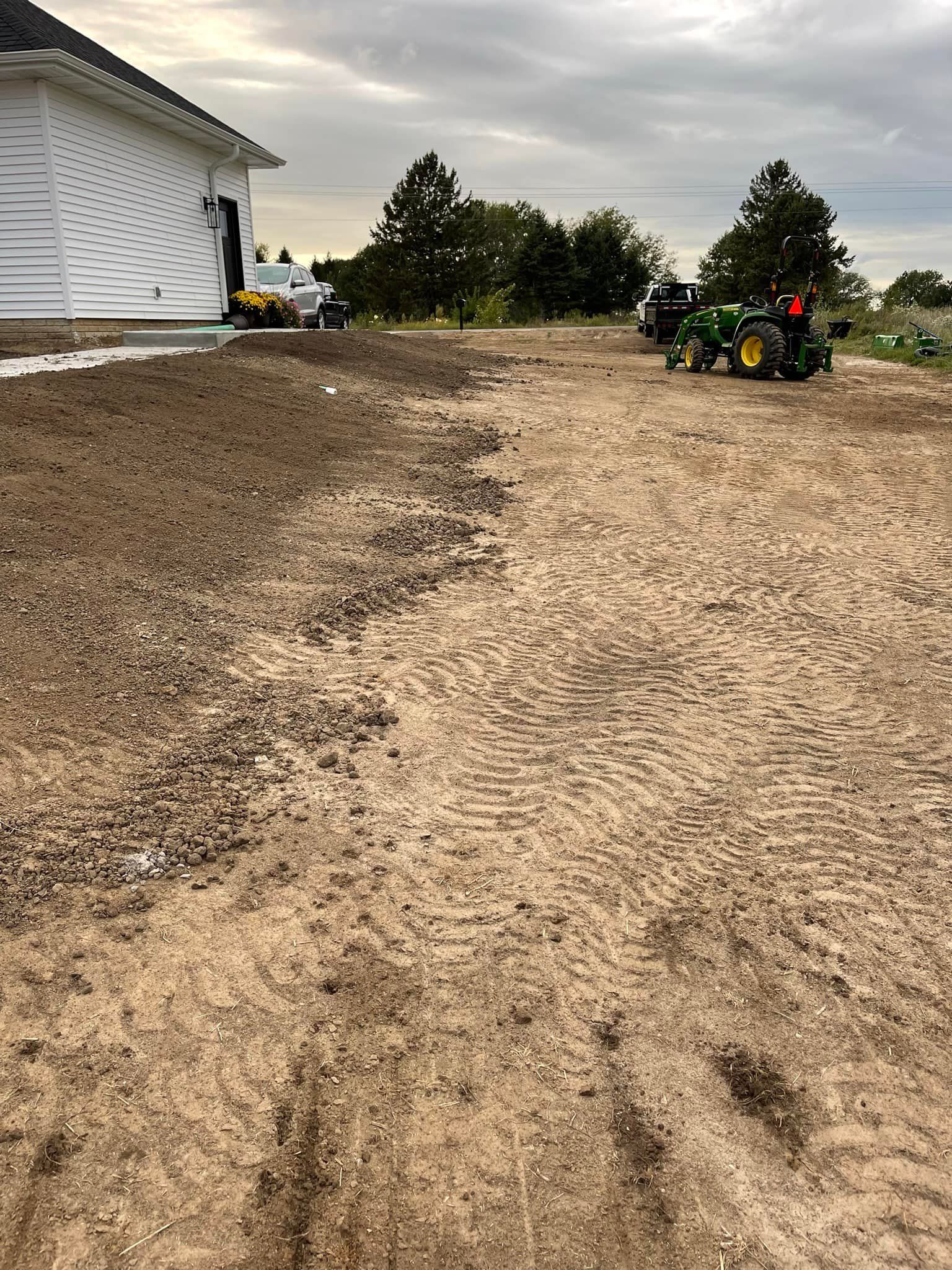 A John Deere tractor working on a dirt yard next to a house under a cloudy sky.