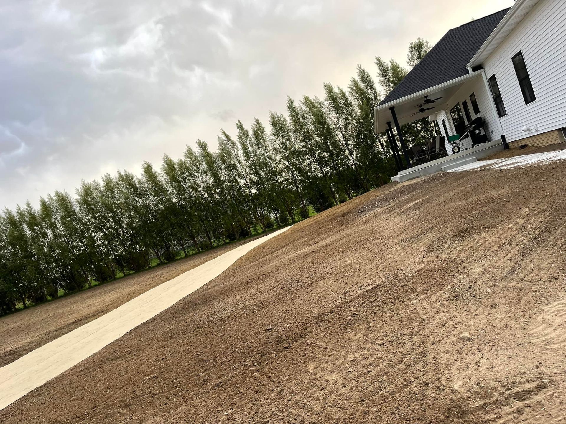 A house with a white exterior and a dark roof. A concret from the house, and rows of trees line the property. Overcast sky.