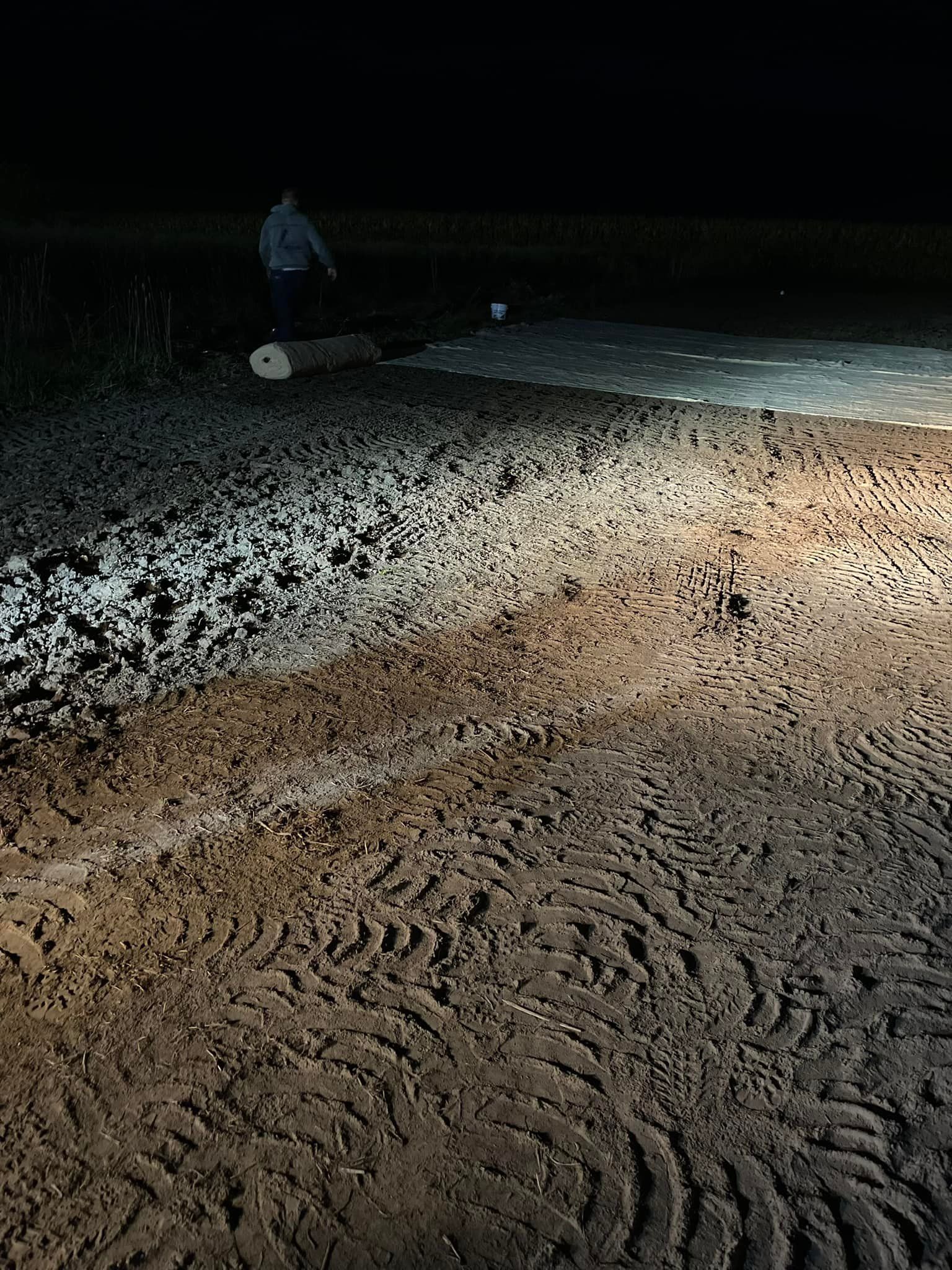 Night scene. Headlights illuminate a muddy road with tire tracks. A person stands in the distance.