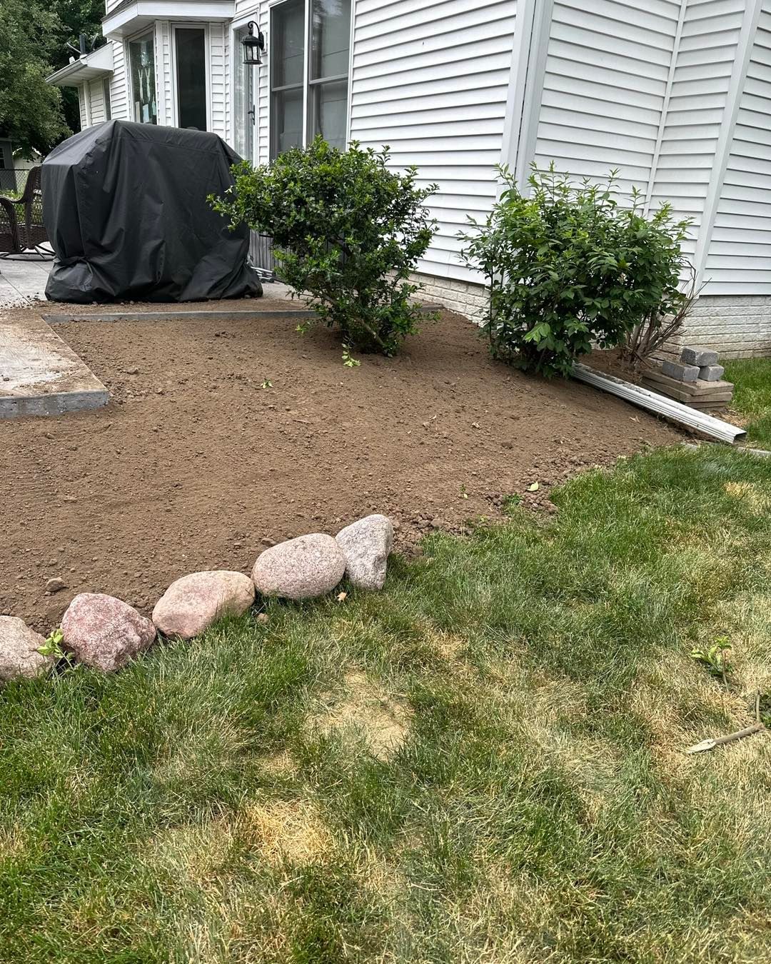 Backyard with dirt patch, bushes, rocks bordering grass, and a covered grill.