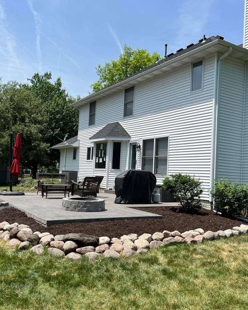 Backyard patio with fire pit, grill, and seating next to a two-story white house with landscaping.