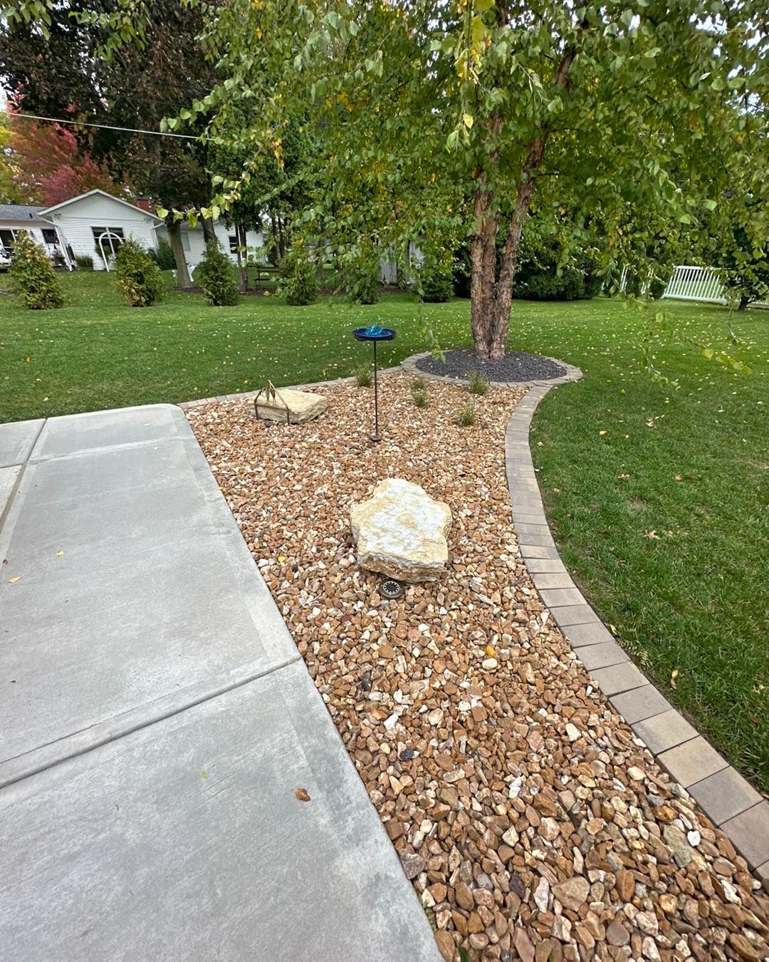 A gravel-filled landscaping bed with a tree, bordered by a concrete walkway and lawn.
