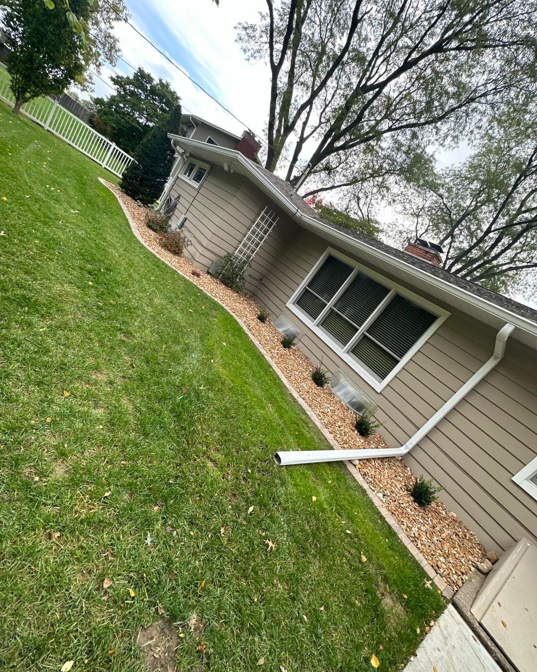 Tan house with a green lawn, gravel landscaping, and a white fence on a sloped yard.