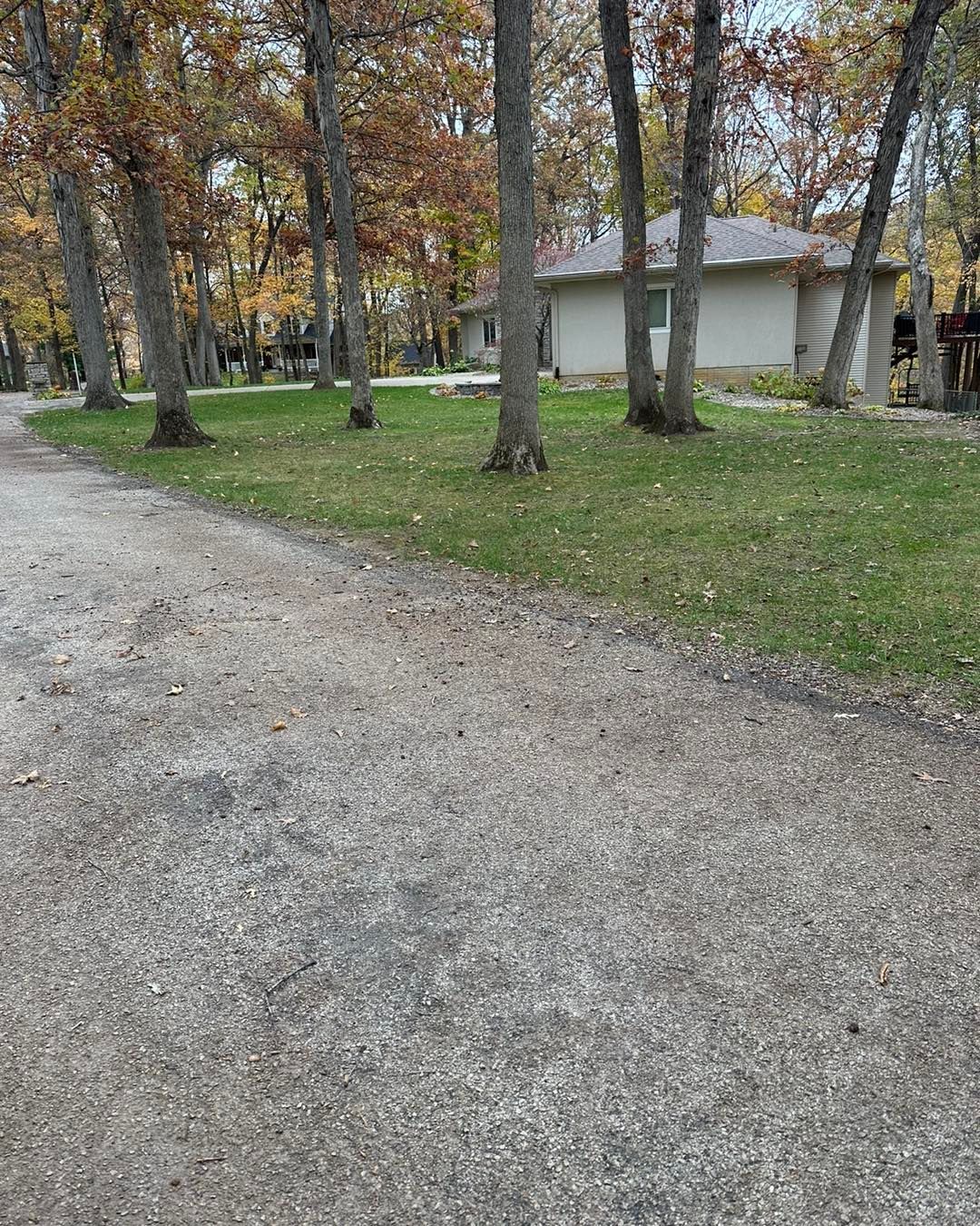 Gravel driveway leads to a one-story beige house surrounded by trees with fall foliage.