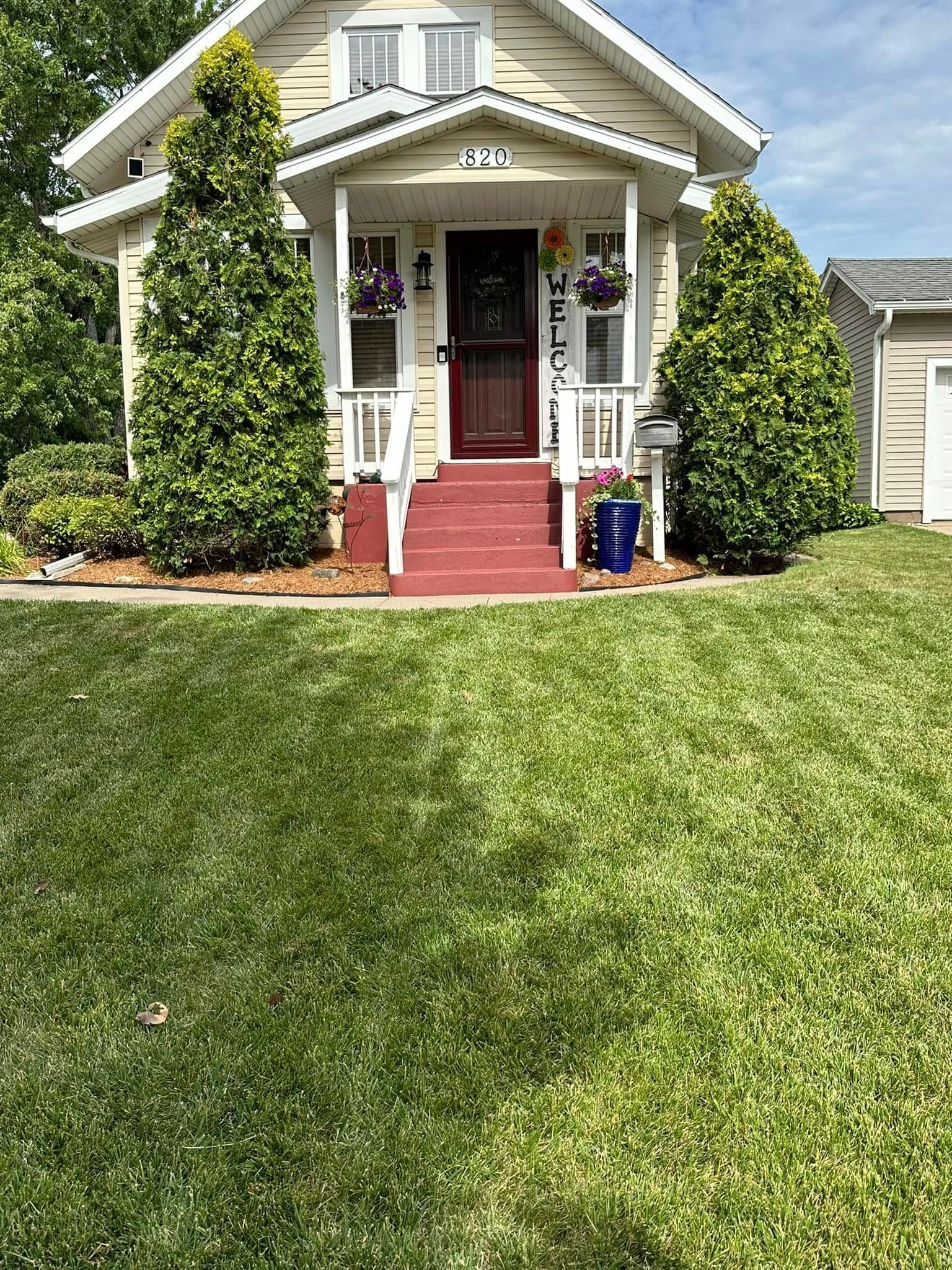 A small house with a porch, red steps, and a well-manicured lawn. Two tall green trees frame the entrance.
