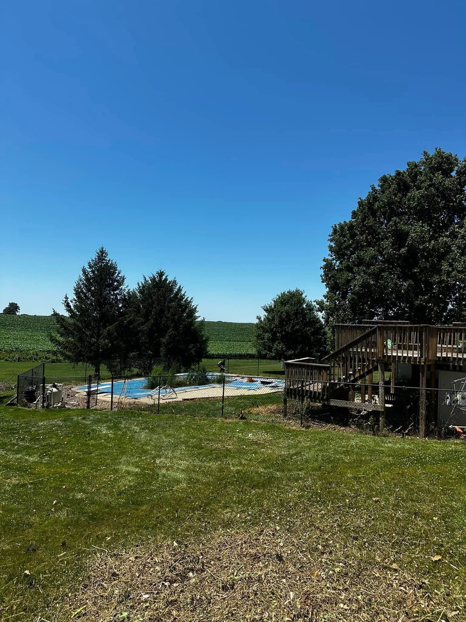 Backyard with a pool, trees, and wooden deck under a bright blue sky. Green grass in the foreground.