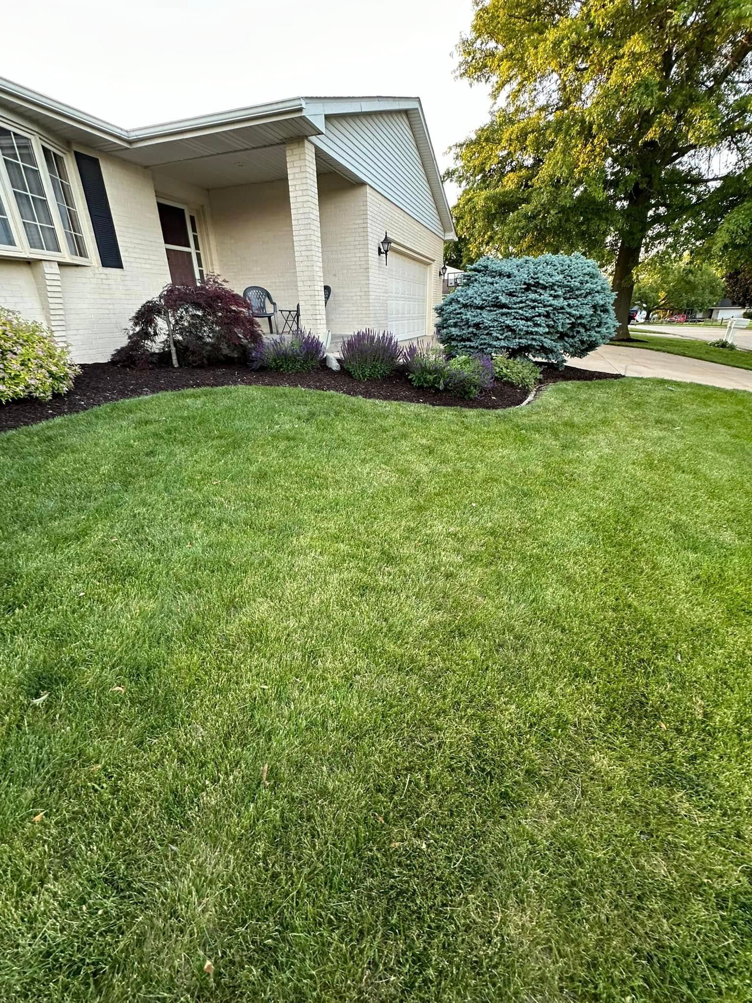 Green lawn in front of a beige house with a garden bed and trees.