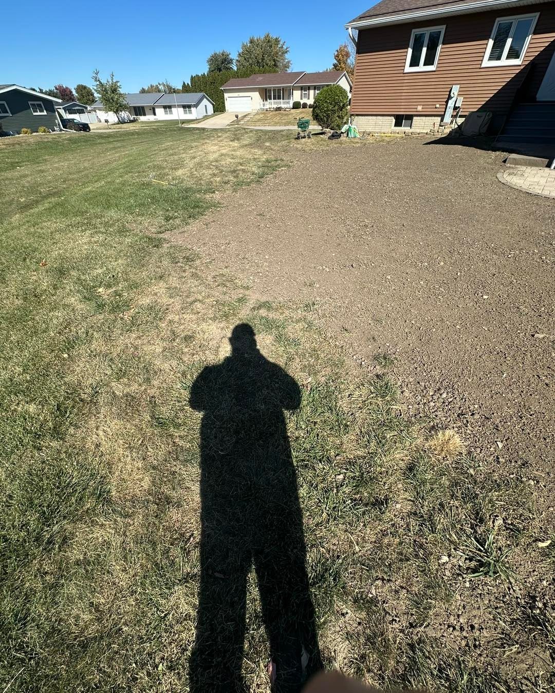 Person's shadow on dry grass and soil, houses in the background on a sunny day.