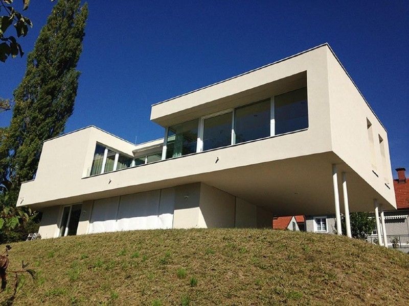 Ein modernes, minimalistisches Haus mit weißen Außenwänden und großen Fenstern, das auf einem grasbewachsenen Hügel 