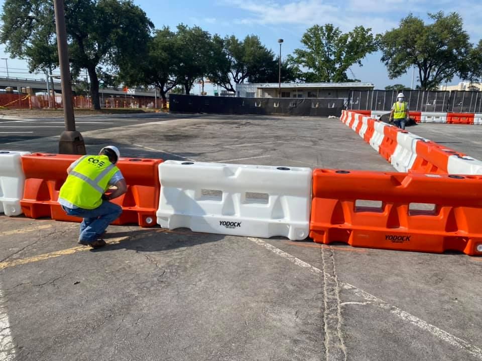 A man in a yellow vest is kneeling next to a row of orange and white water filled barriers.