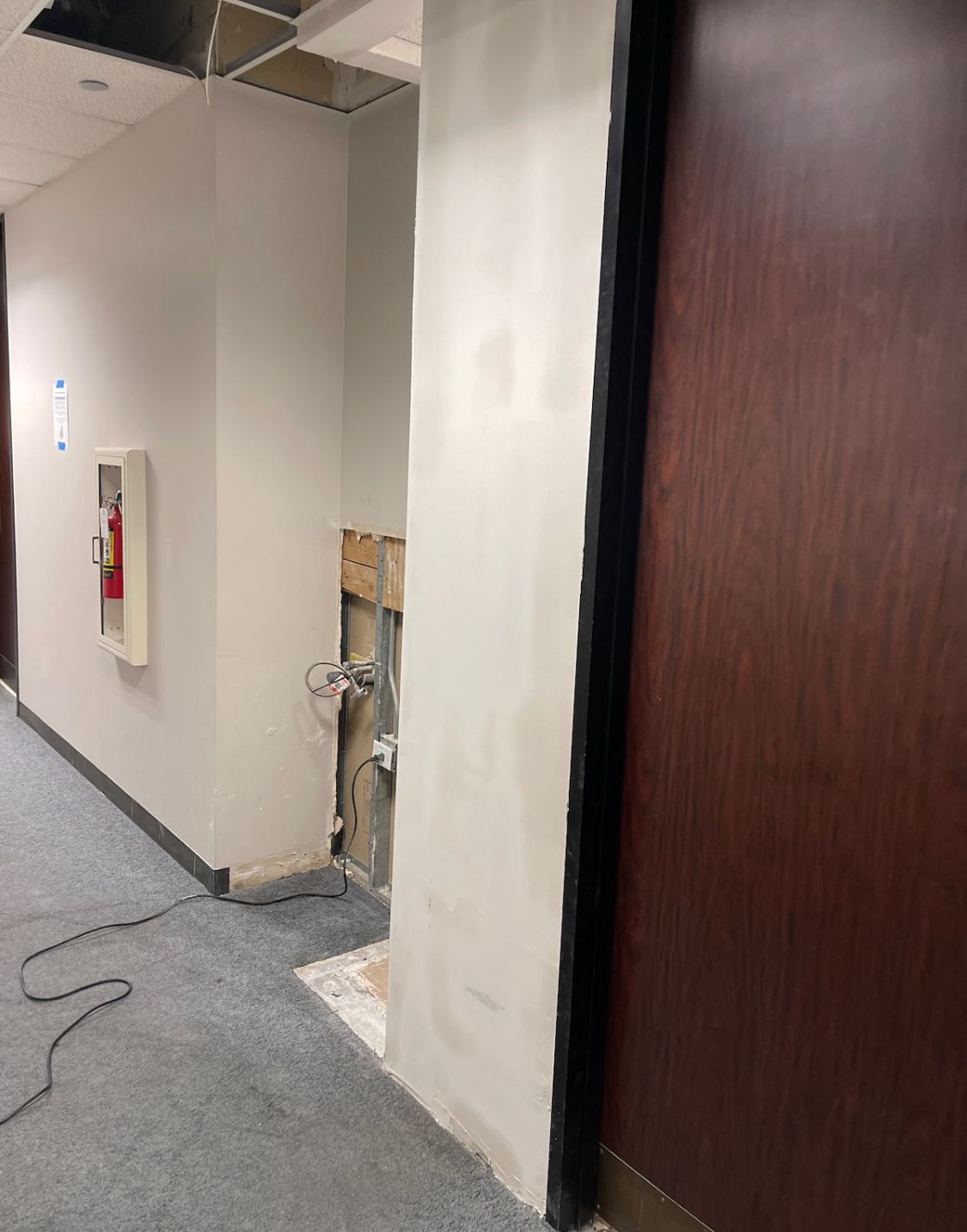 A hallway with a hole in the wall and a fire extinguisher on the wall.