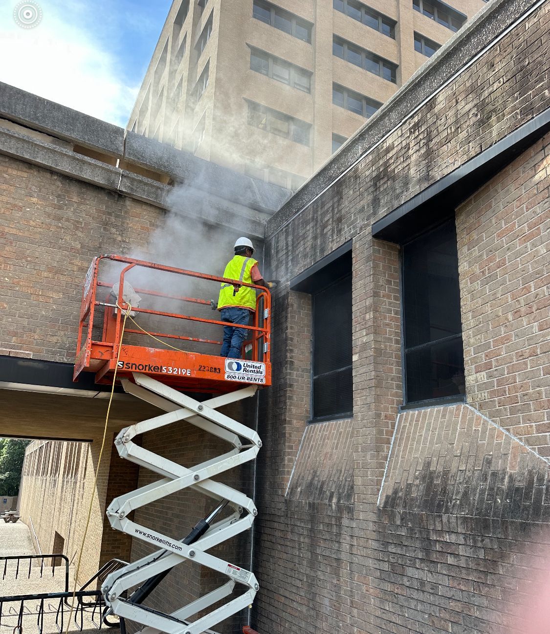 A man is standing on a scissor lift spraying water on a brick building.