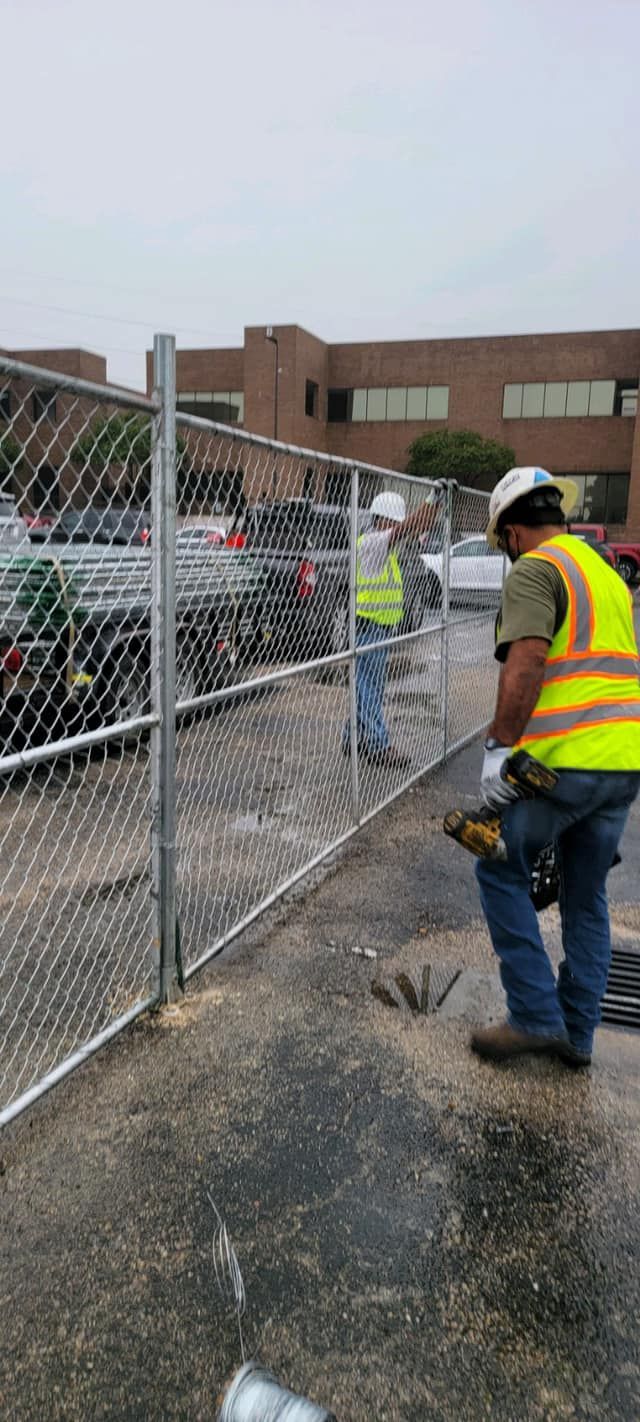 A man in a yellow vest is working on a chain link fence.