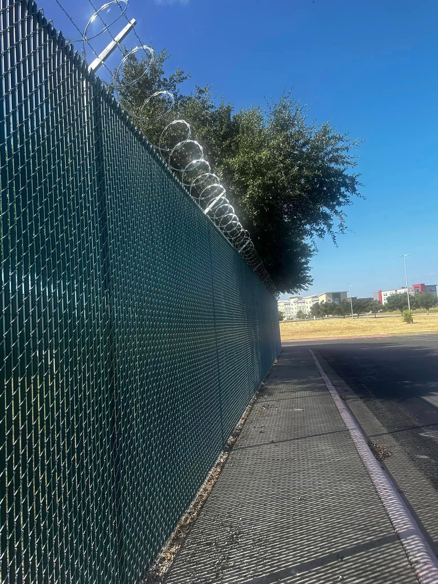 A green fence along a sidewalk with a barbed wire fence