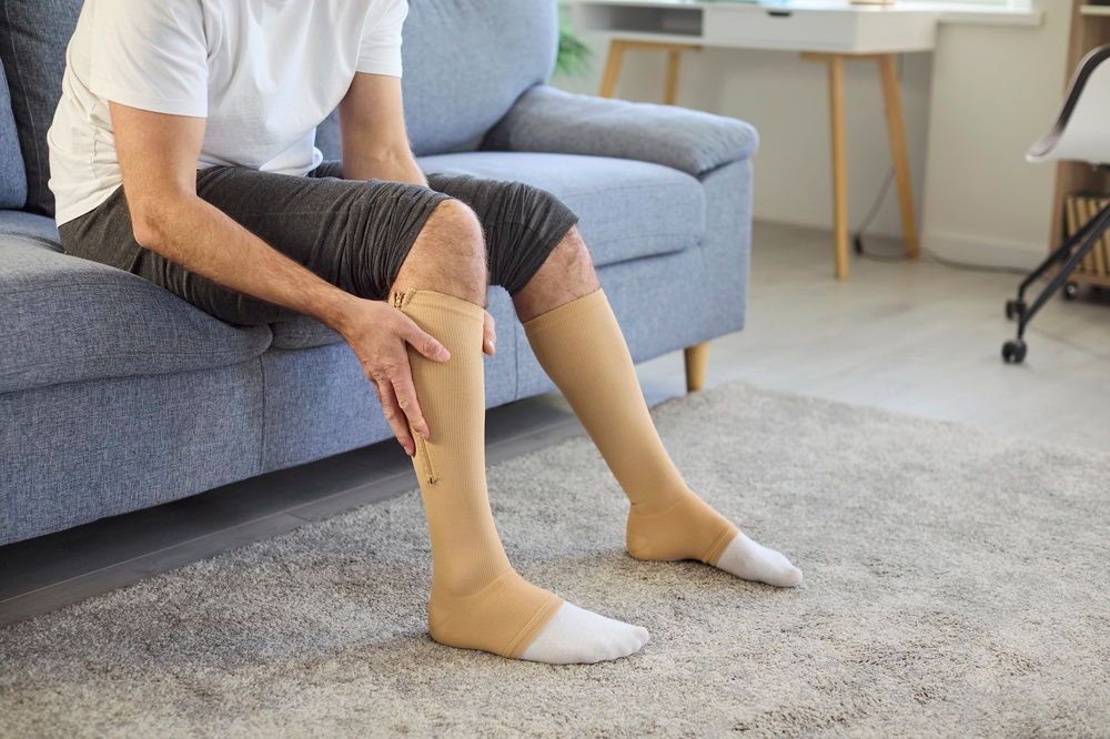 Man seated on a sofa wearing compression socks, touching his leg. Indoors, neutral tones.