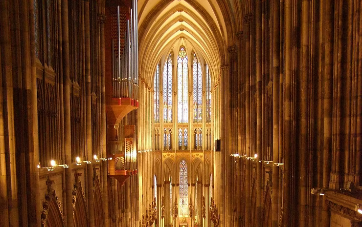 Interior view of a Gothic cathedral, with rows of columns and a large stained-glass window at the end, illuminated.