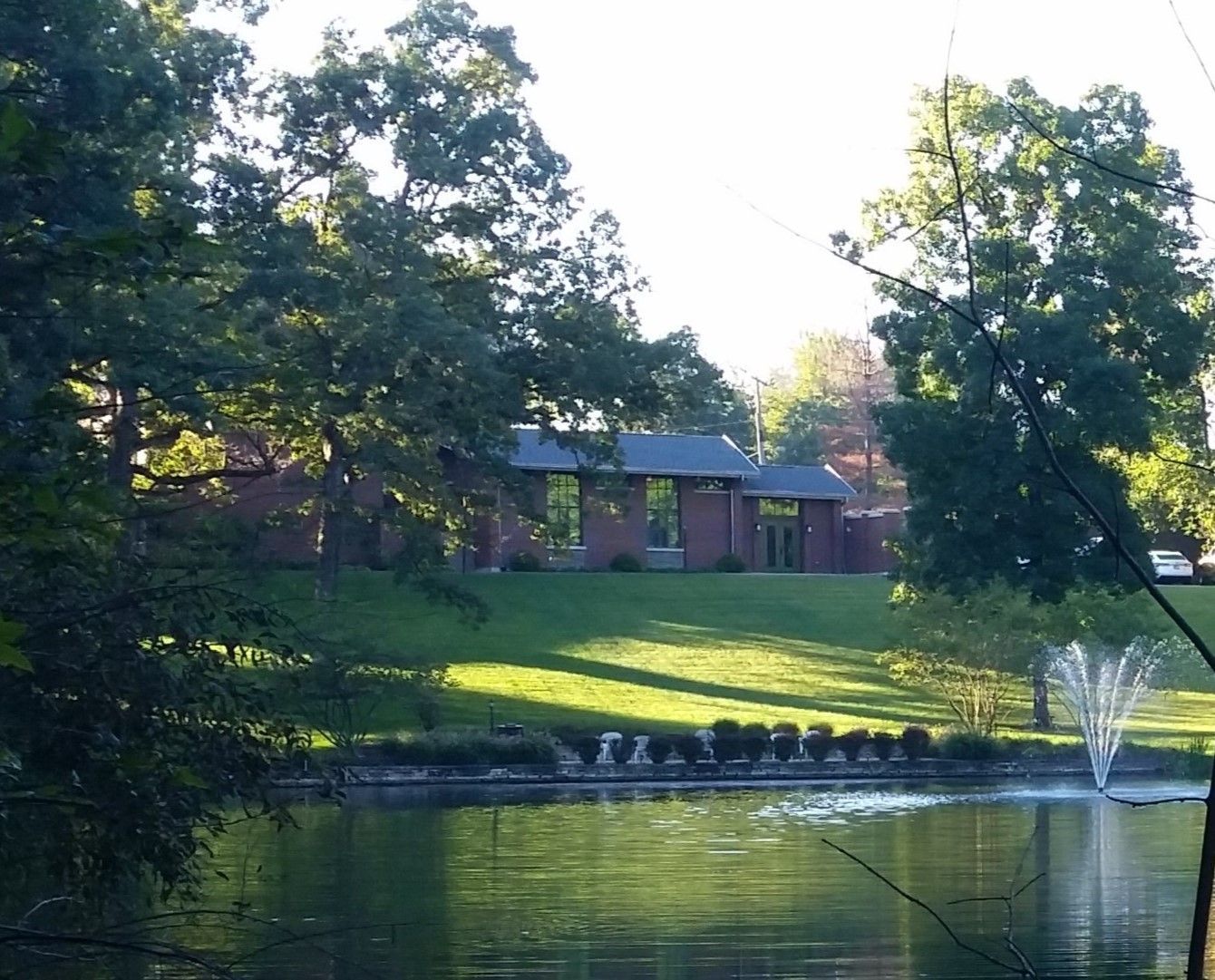 Pond in park with brick building, trees, and fountain under a sunny sky.