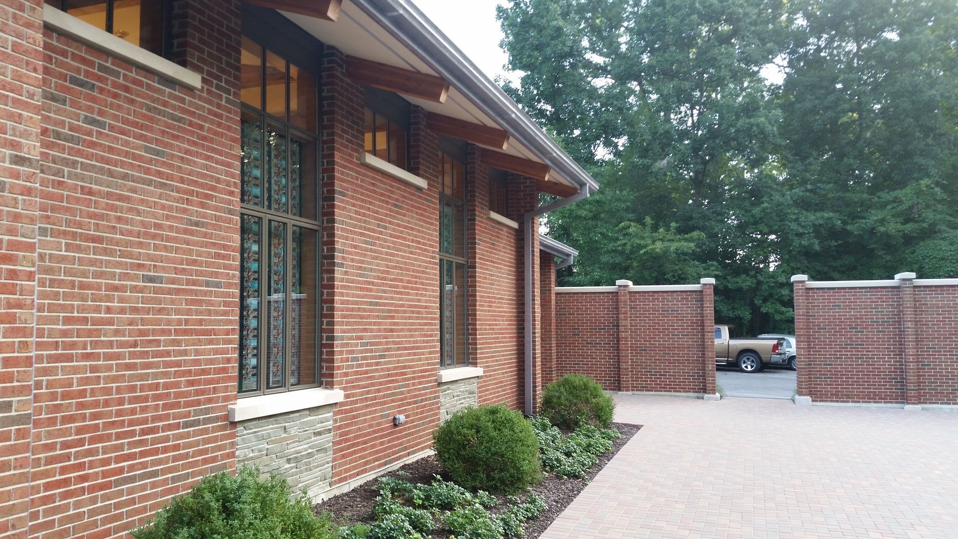 Red brick building with windows, wood trim, and a brick paved walkway next to manicured bushes and a wooden fence.