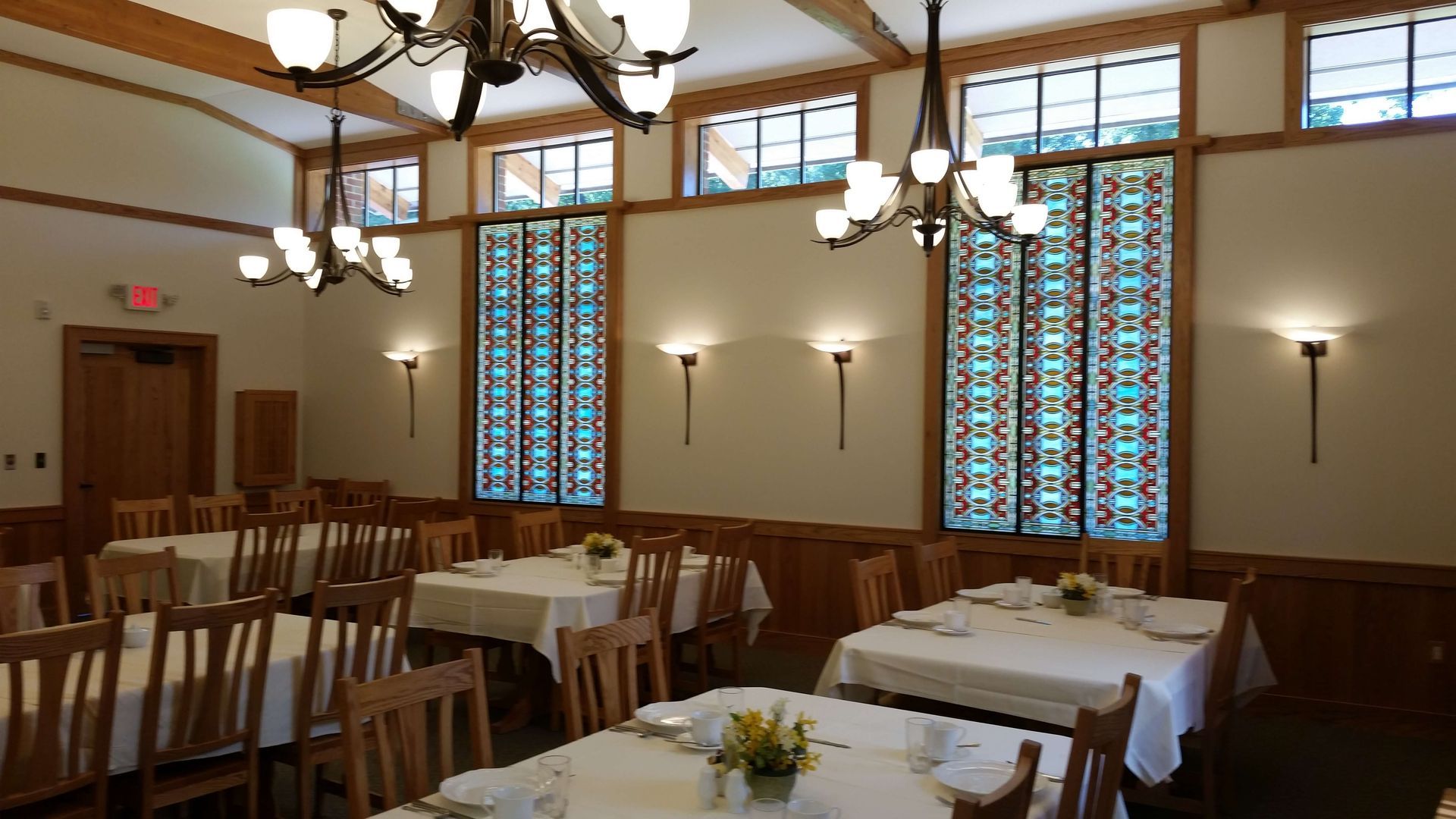 Dining hall with tables set for a meal, wood furniture, chandeliers, sconces, and stained-glass windows.