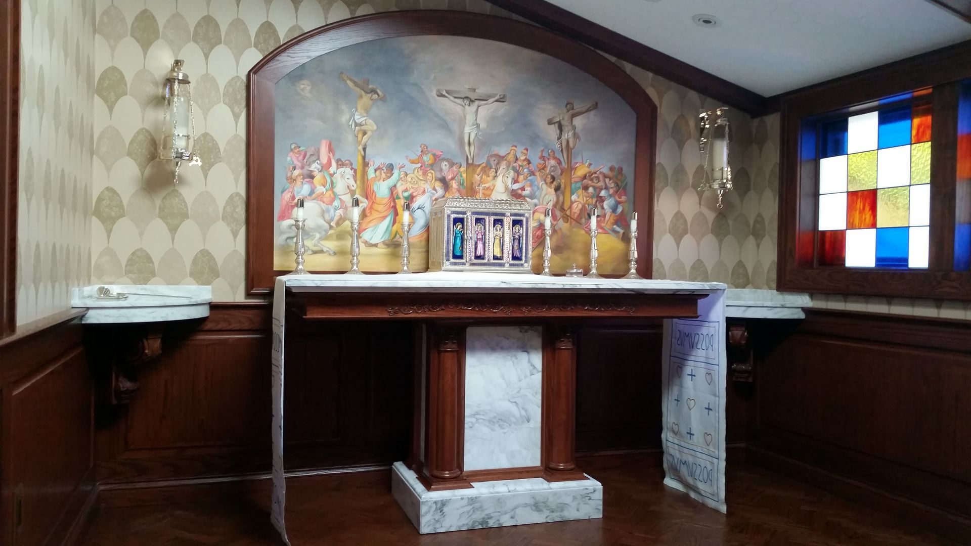 Altar with painting of religious figures, candles, and stained glass window in a chapel.