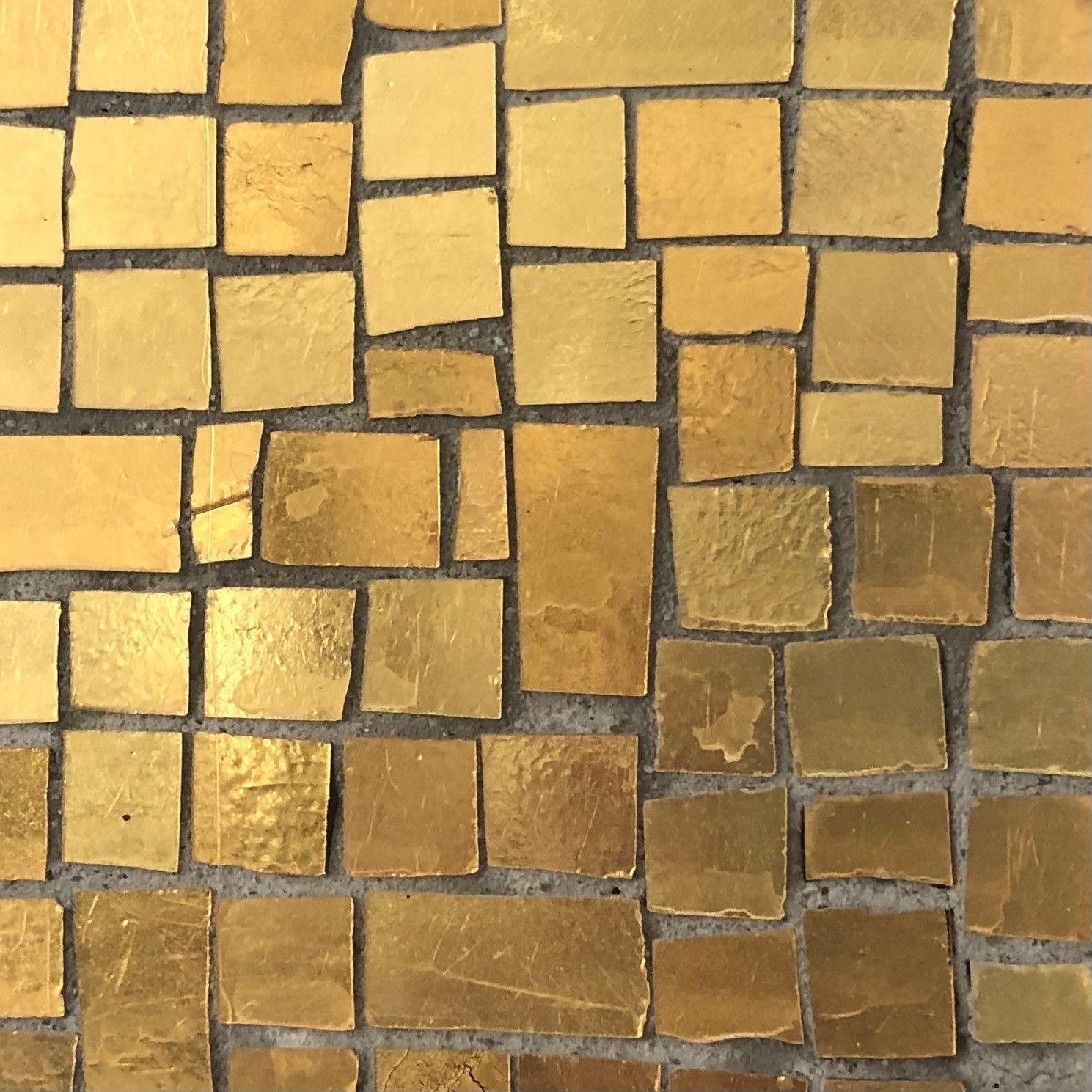 Gold mosaic tile pattern. Squares of varying sizes and tones of gold on a gray surface.