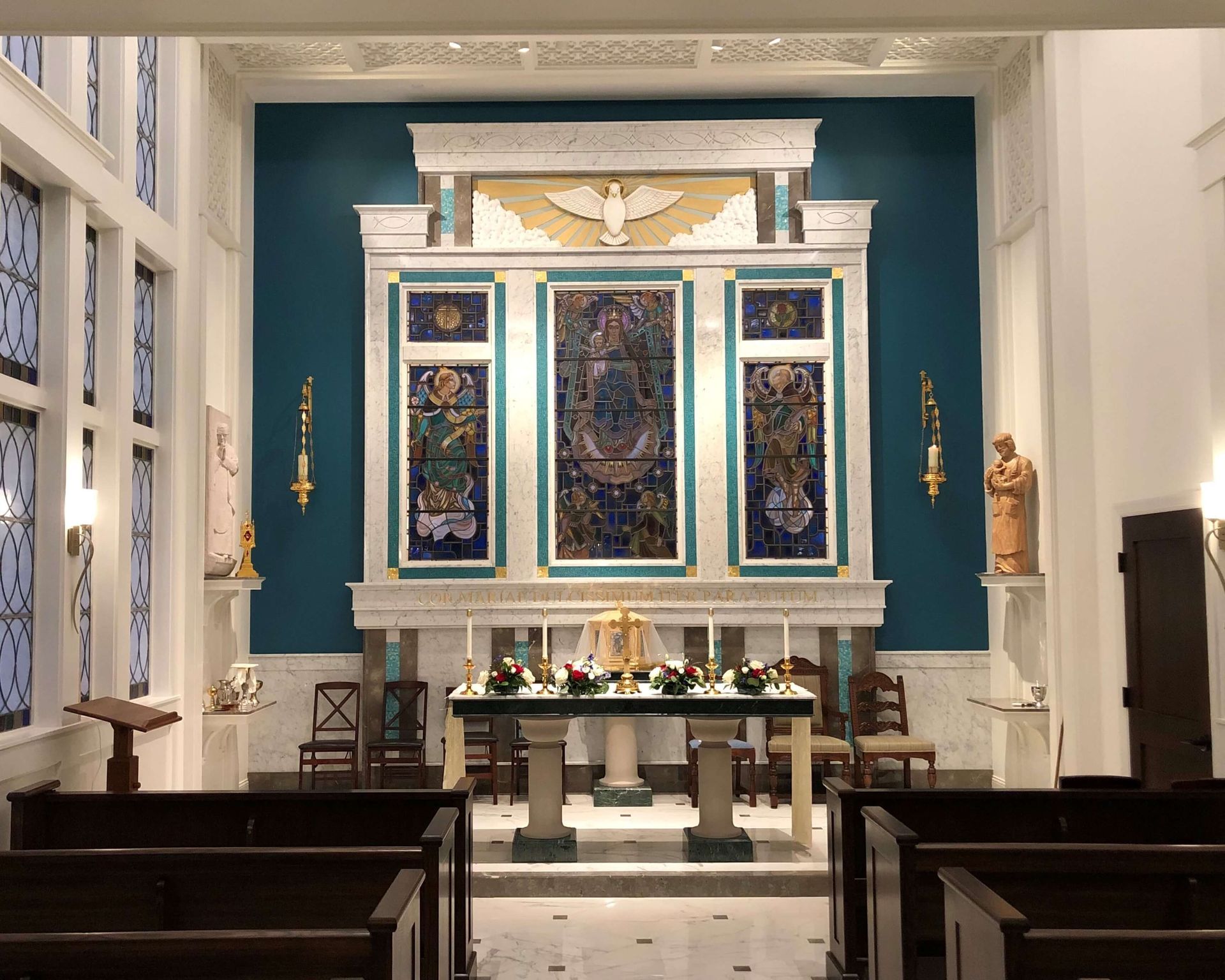 Altar in a church with teal background, stained glass, candles, and a dove emblem.