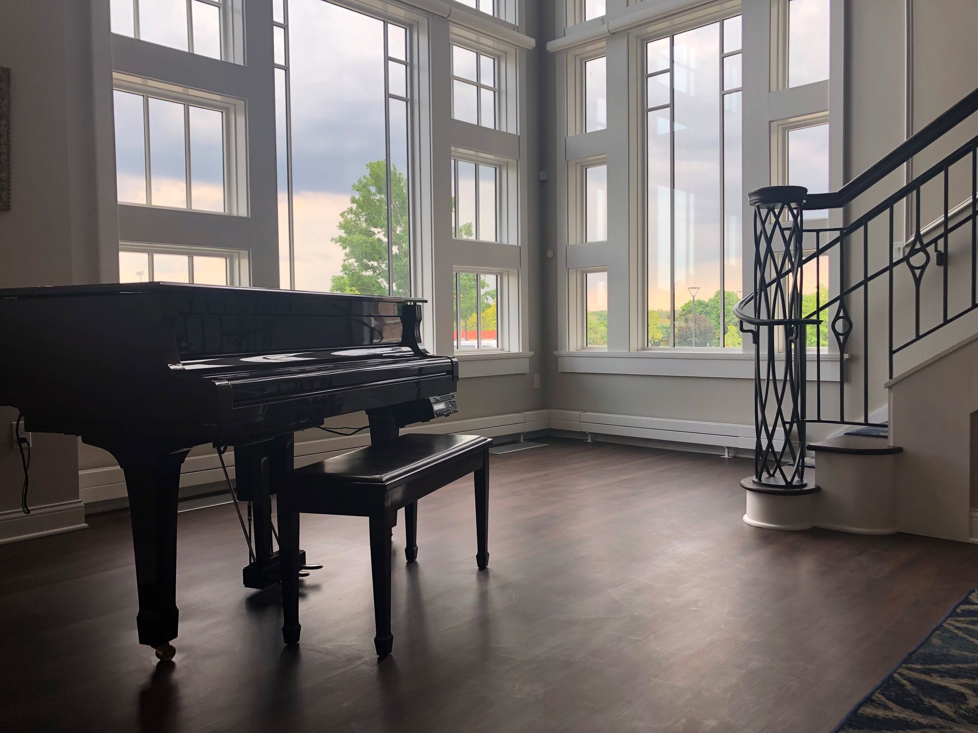 A grand piano sits in a room with large windows, near a staircase.
