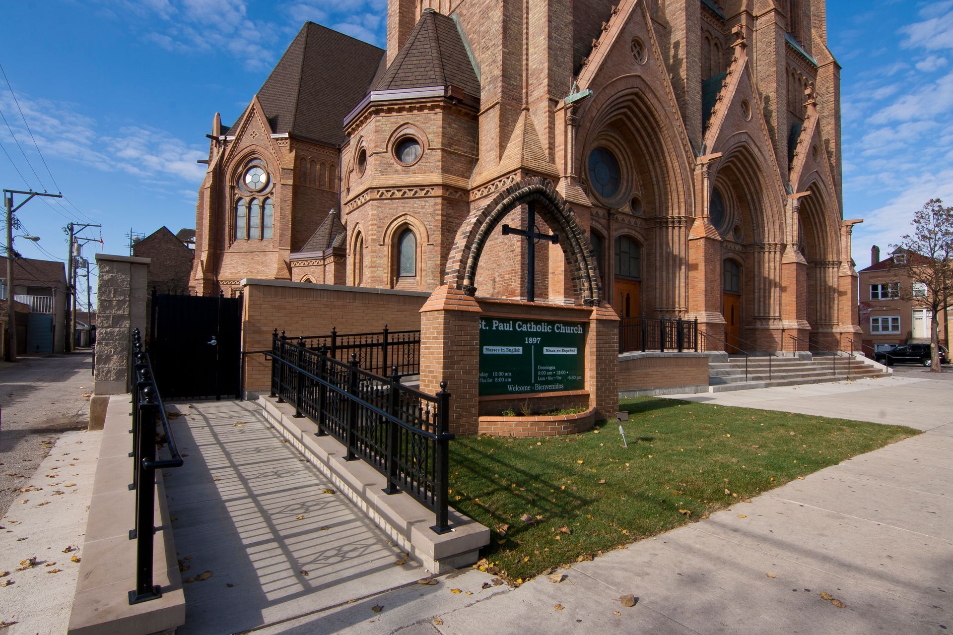 Church building with a ramp leading to the entrance. Sign with cross and green panels, brown brick exterior, blue sky.