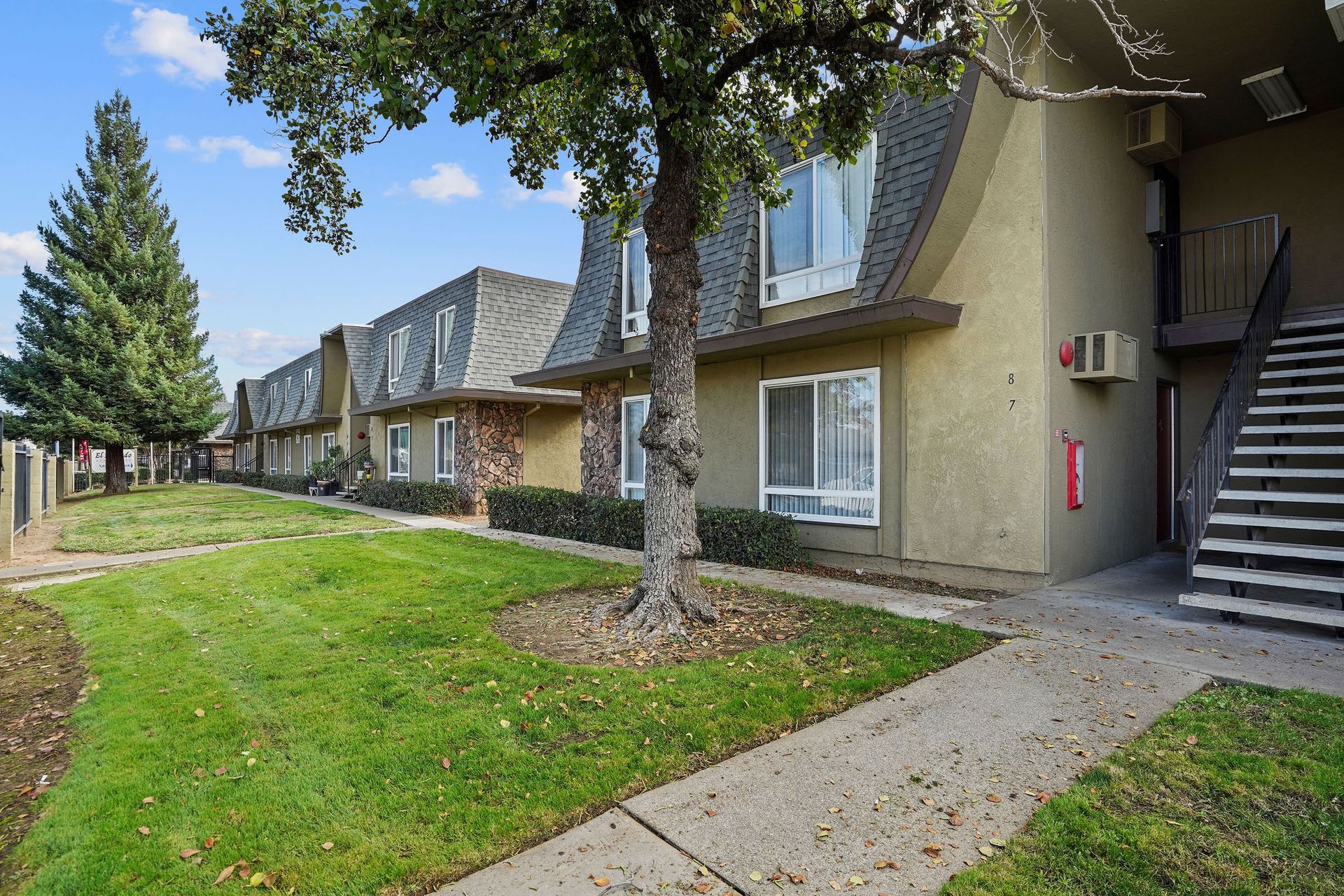 Apartment complex courtyard with trees, grass, and exterior stairways beside beige buildings