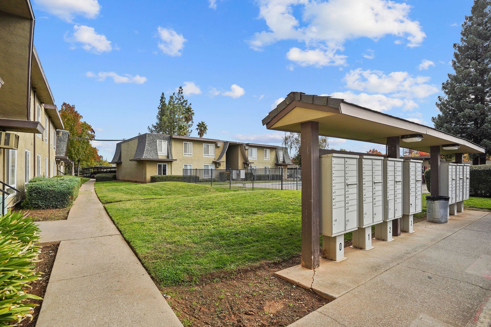 Apartment courtyard with mailbox shelter, sidewalk, and grassy lawn under a blue sky