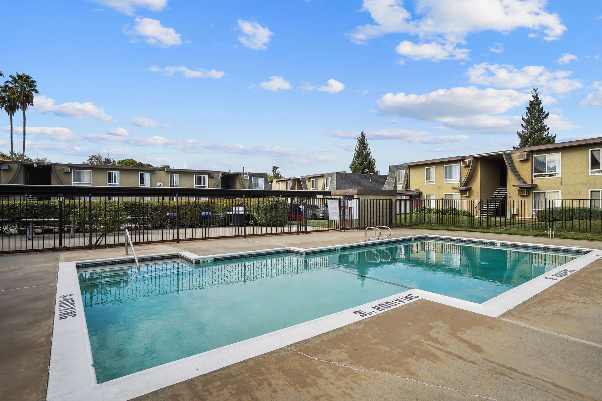 Outdoor apartment complex pool with turquoise water and lounge chairs under a partly cloudy sky