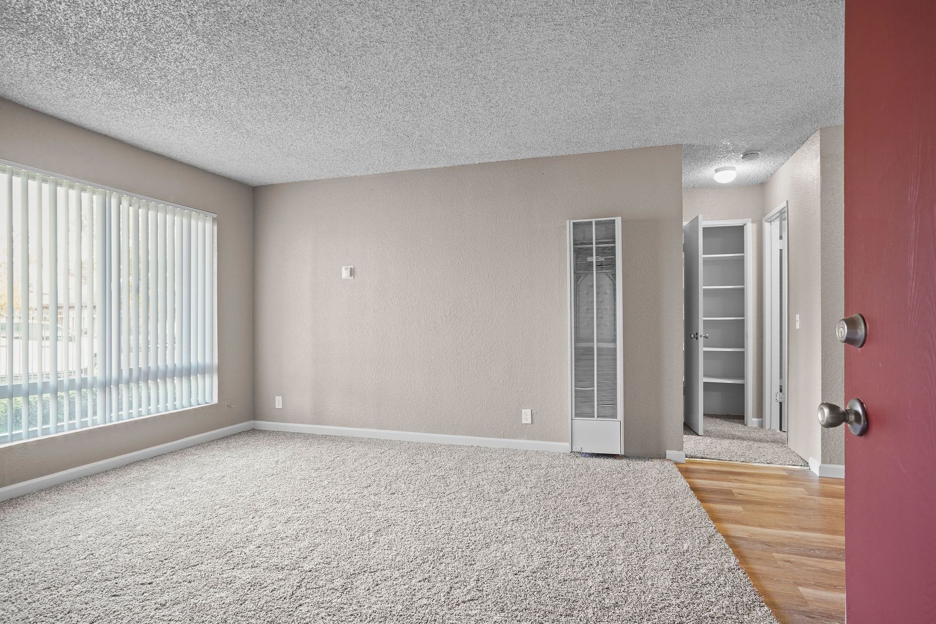 Empty living room with carpet, large window blinds, and hallway to the right.