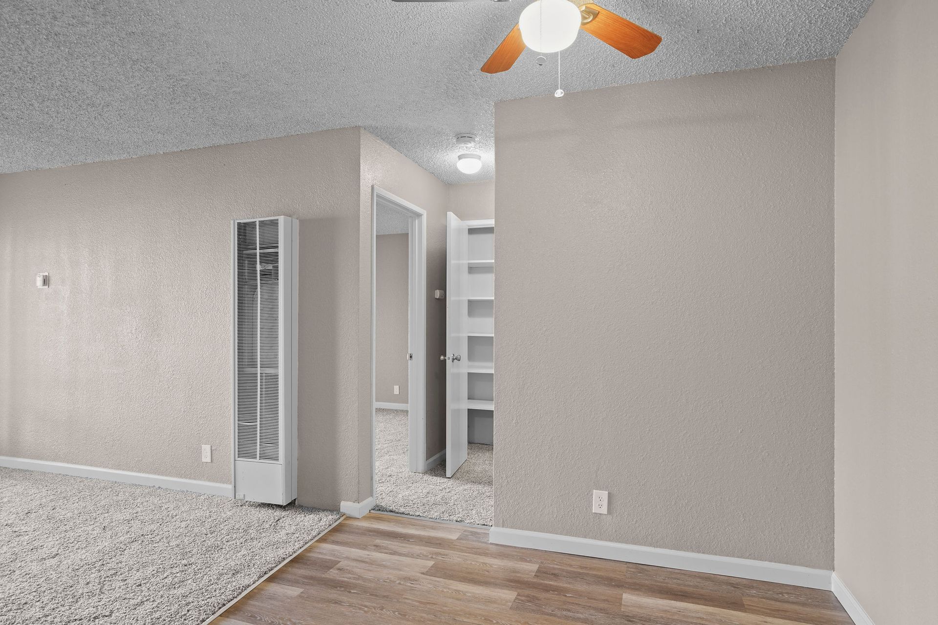 Empty beige room with carpet, wood floor, ceiling fan, and open doorway to a hallway with shelves