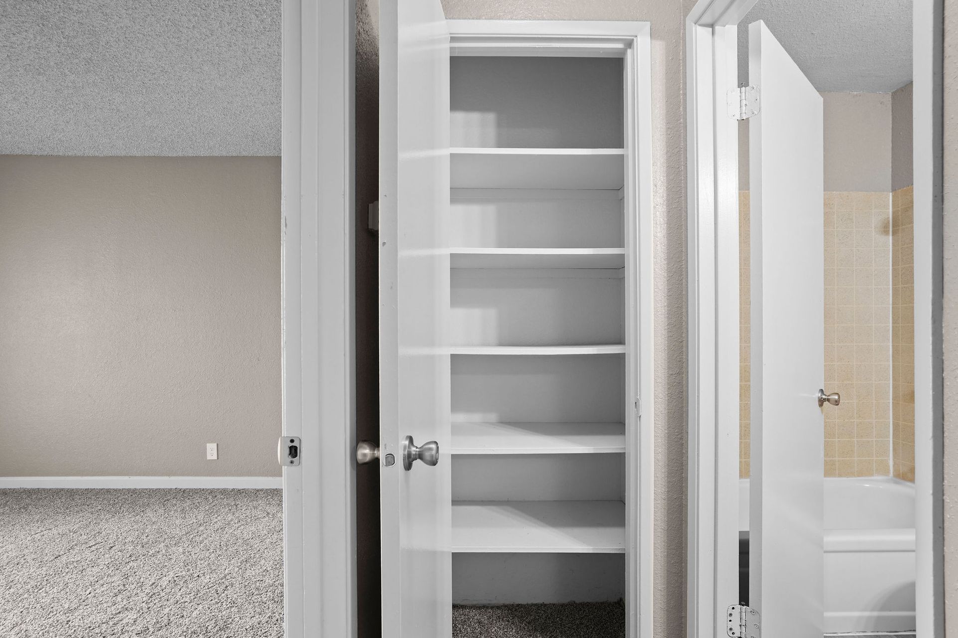 Open closet with white shelves between a carpeted room and bathroom doorway