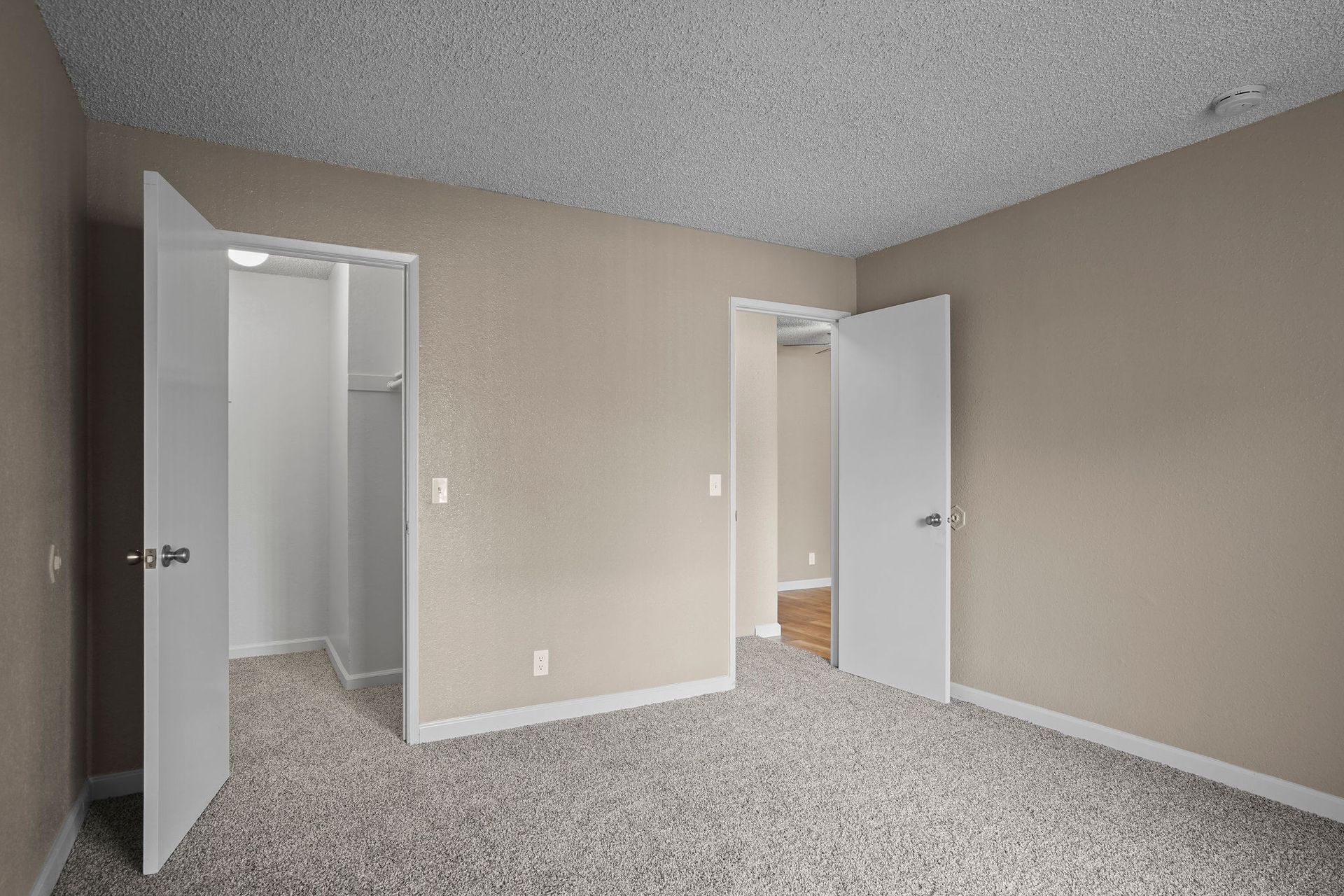 Empty beige carpeted room with open double doors leading to a bathroom and another room