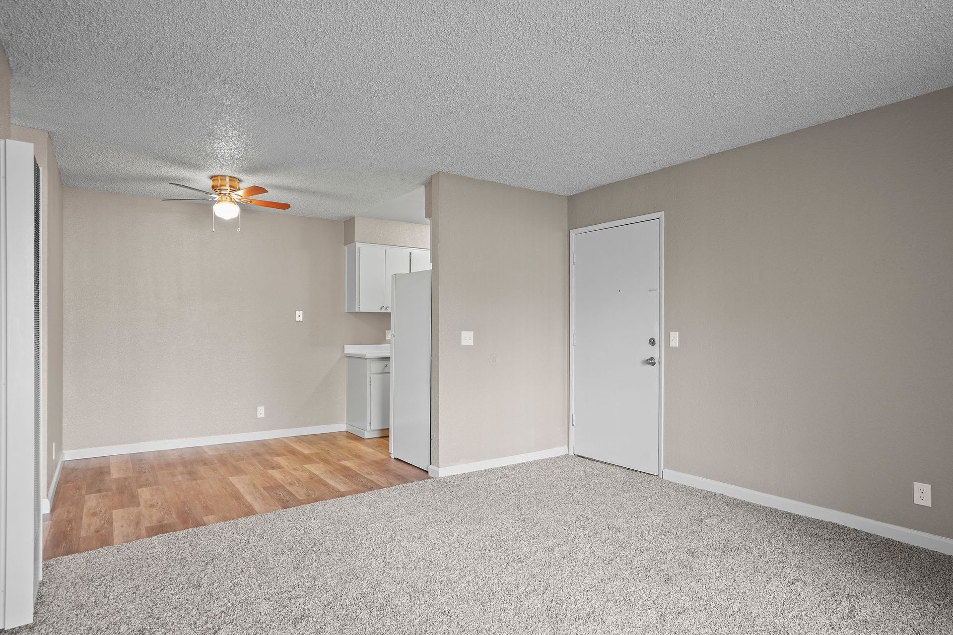 Empty living room with gray carpet, beige walls, and a small kitchen area visible in the back.