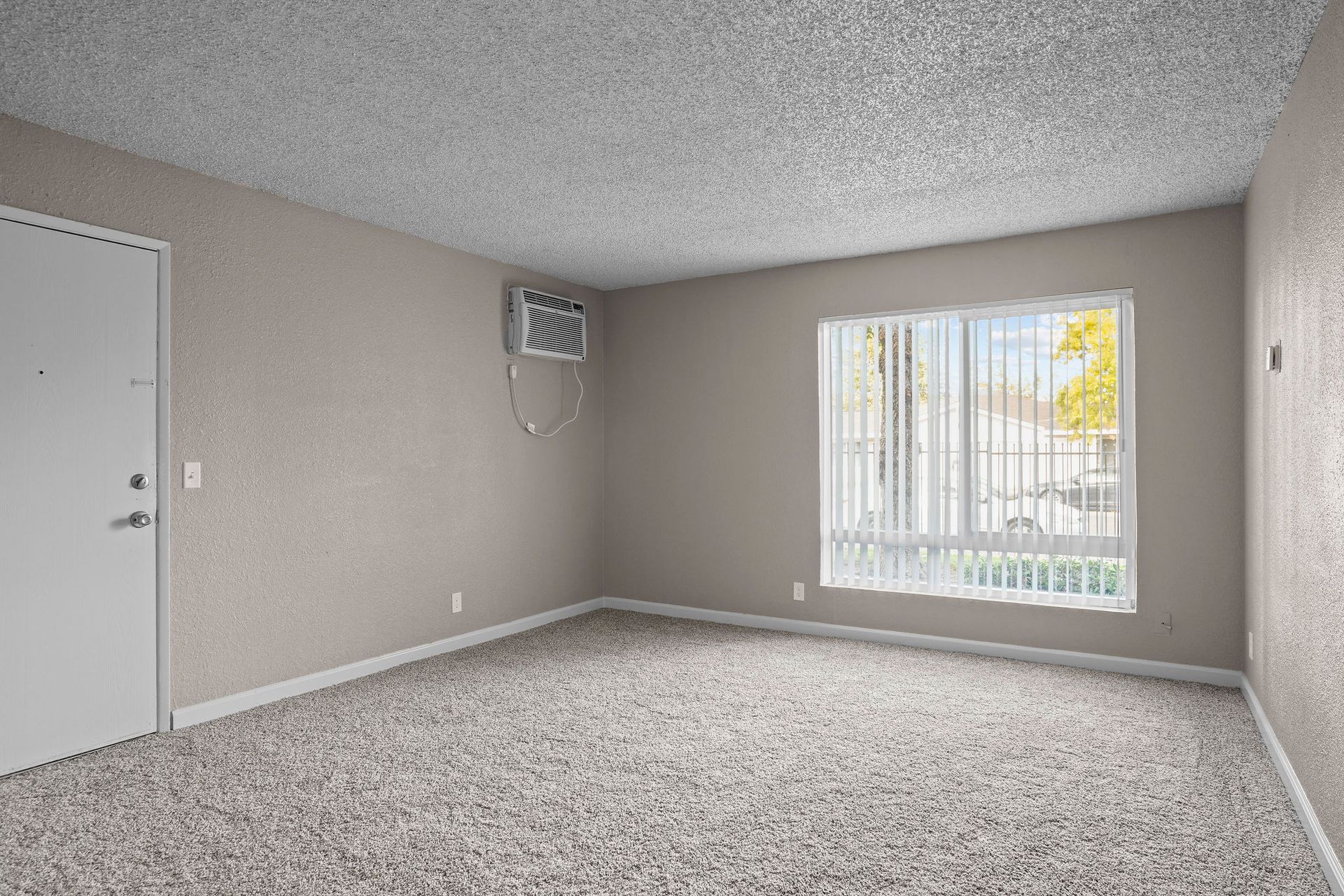 Empty beige living room with carpet, white door, and window with vertical blinds