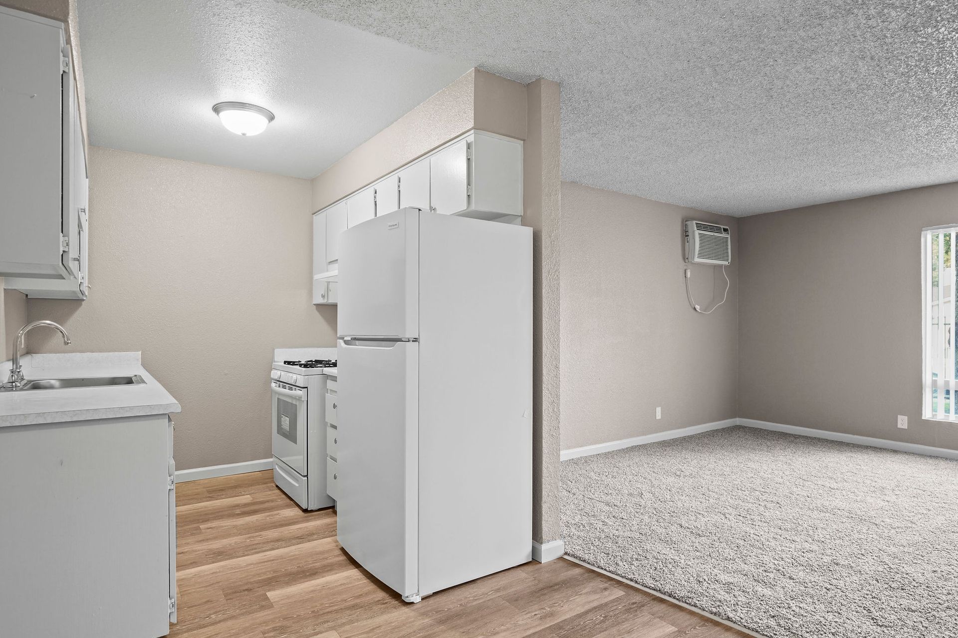 Empty kitchen and carpeted living area in a beige apartment, with white appliances and wood flooring.