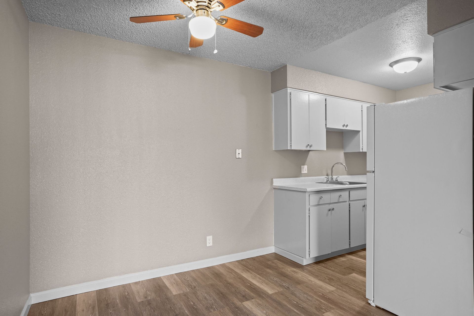 Empty apartment kitchen with gray cabinets, white refrigerator, wood floor, and ceiling fan.