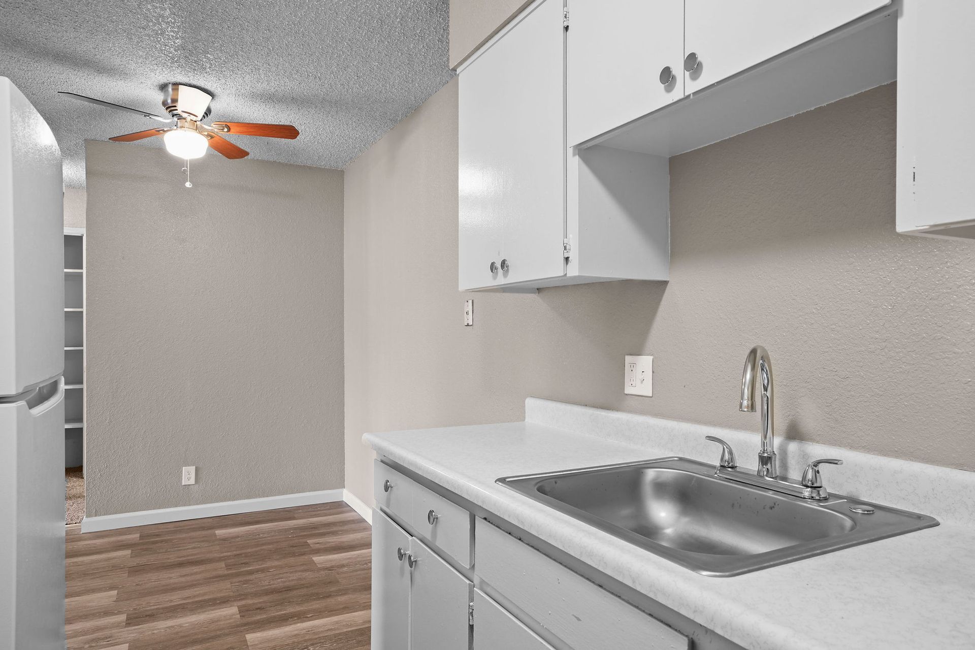 Small kitchen with white cabinets, gray countertop, sink, and ceiling fan, opening into a hallway.