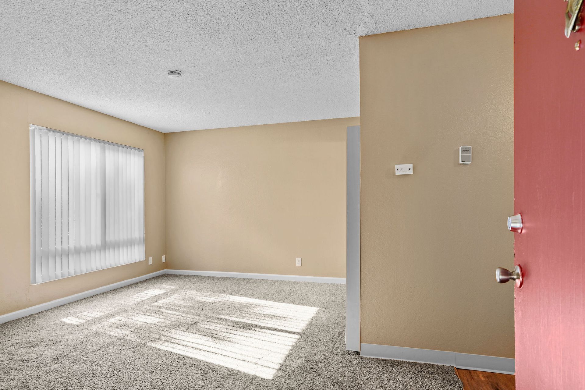 Empty beige living room with carpet, sunlight from a window, and a red doorway on the right