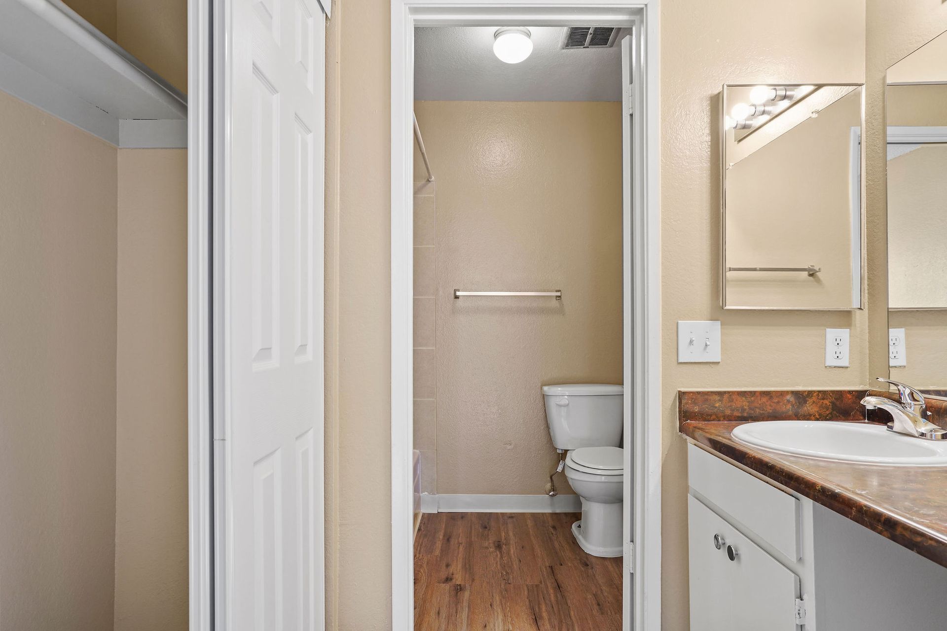 Narrow bathroom hallway with a toilet and vanity, beige walls, and wood flooring.