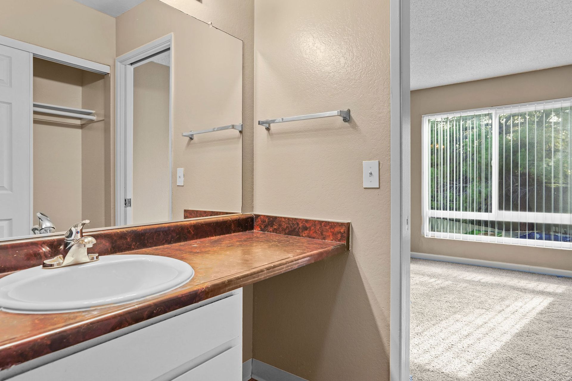 Bathroom vanity with granite countertop and sink, looking into a carpeted bedroom with a window and vertical blinds